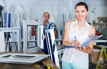 Young cheerful positive smiling woman manager standing with clipboard in workshop for production of plastic windows