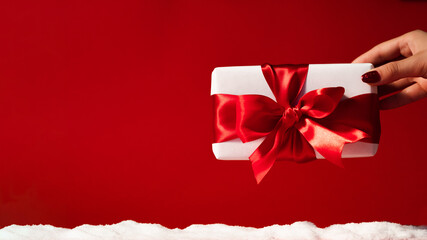 Christmas gift. Winter holidays greeting. Female hand holding wrapped present in white box with ribbon bow isolated on red copy space decorative background with snow pile. Special day congratulation.