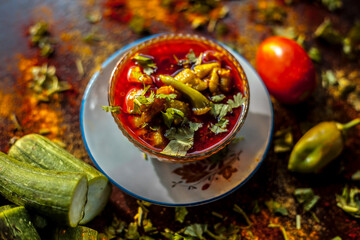 Close up shot of fresh Indian Lunchtime dish luffa or Galka nu Shaak in a glass container along with all its ingredients on a rust-colored surface.