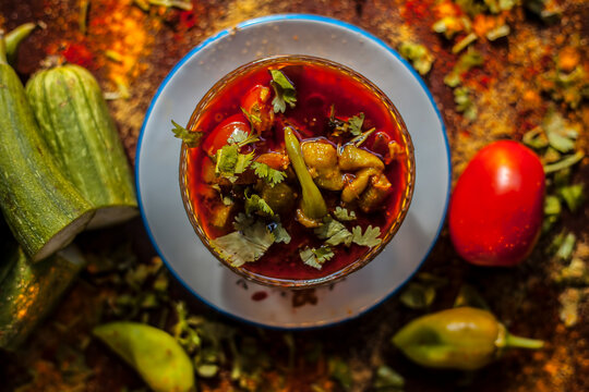 Close up shot of fresh Indian Lunchtime dish luffa or Galka nu Shaak in a glass container along with all its ingredients on a rust-colored surface.