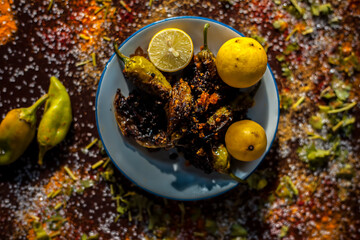 Close up shot of Classic Bharela Marcha or Stuffed fried chilies in a glass plate along with lemons and all the spices needed for preparing it on a brown wooden surface.