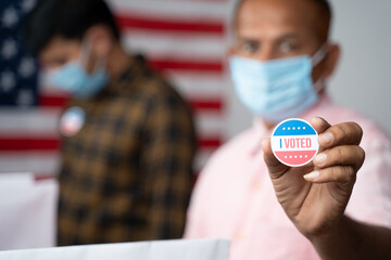 Close up of Hands, Man in medical mask showing I voted Sticker at polling booth with US flag as...