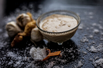 Shot of freshly ground garlic and ginger paste in a glass bowl on a black surface along with raw ginger, and some garlic with salt on sprinkle on the surface. 