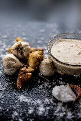 Shot of freshly ground garlic and ginger paste in a glass bowl on a black surface along with raw ginger, and some garlic with salt on sprinkle on the surface. 