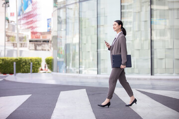 Asian woman with smartphone walking against street blurred building background, Fashion business photo of beautiful girl in casual suite with smart phone. 