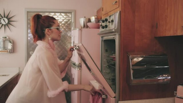 A young woman standing in a retro styled kitchen, hears that her food is ready and turns to the oven.