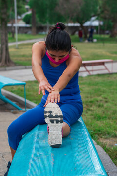 mujer joven con cubrebocas realizando estiramiento sentada en banca azul