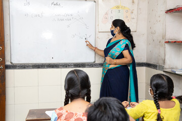 Indian teacher and students wearing face masks maintaining social distancing study in classroom back at school during covid19 pandemic.