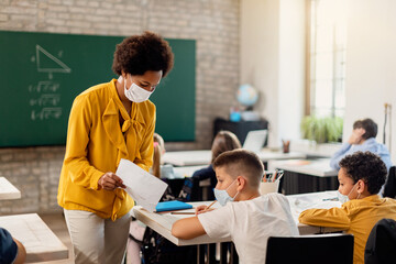Black teacher and elementary student with face masks examining test result in the classroom.