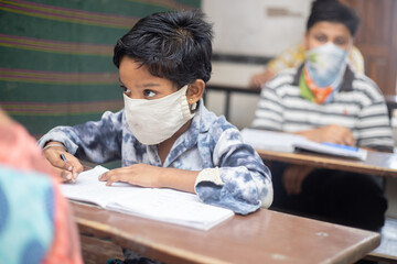 Indian boys students studying In classroom wearing mask maintaining social distancing looks at camera, school reopen during covid19 pandemic, new normal. selective focus