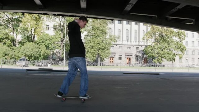 Professional skateboarder guy in dark clothing performs trick on skateboard. Teenager rides on front wheels of skateboard, performing balancing tricks on site of skate park under an iron bridge.