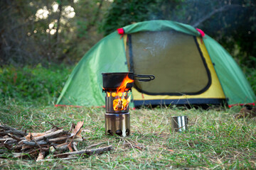 food is cooked on a camping wood stove. green tent on the grass at the background, firewoods and steel mug near the wood stove