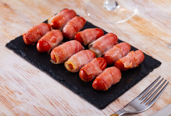 Grilled small smoky cocktail wieners wrapped in bacon strips on slate serving board