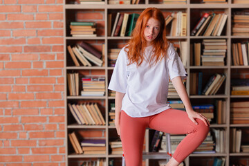 Redhead female college student taking book from shelf in library. Stands on the stairs to get a book from the top. Copy space.