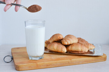 A hand pours cocoa into milk with a spoon against the background of a heap of croissants.