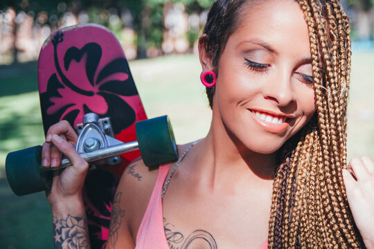 Young woman with tattoos and braids with her longboard smiling in a park during a sunny day