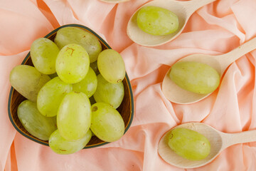 Green grapes in wooden spoons and bowl on pink textile background, flat lay.
