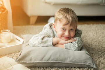 A beautiful little boy in a knitted sweater is reading by the window with a mug of hot tea. Cozy. Autumn.