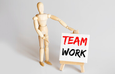 Wooden man shows with a hand to white easel with text TEAM WORK