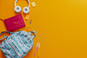 School supplies flat lay on the orange background. Blue backpack, white headphones, notebook and pens.