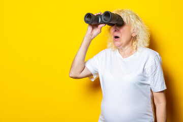 Old woman with a surprised face in a white t-shirt looks through binoculars on a yellow background