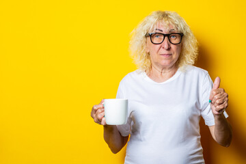 Old woman in white t-shirt and glasses holds a cup of tea on a yellow background