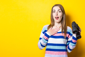 Young woman with a surprised face holds binoculars on a yellow background. Banner