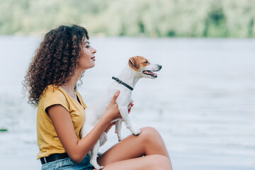 Side view of curly woman in summer outfit holding jack russell terrier dog and looking away while sitting near lake