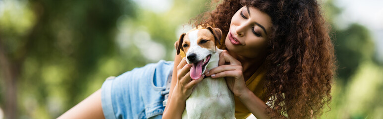 horizontal orientation of curly woman cuddling jack russell terrier dog while resting in park