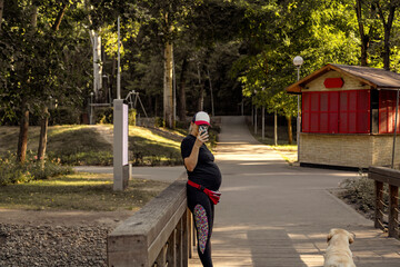 A young pregnant woman in sportswear photographs her dog on the phone. City park, sunny warm day.
shoots on the phone.