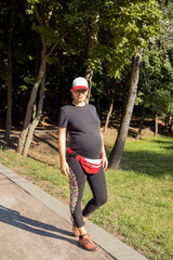 Portrait of a young pregnant woman in sportswear and a baseball cap on a walk in the city park. Close-up, selective focus.