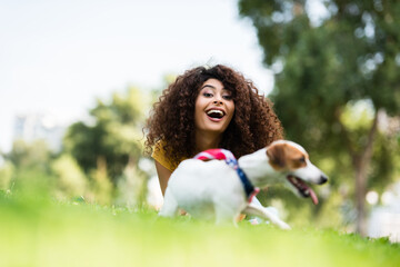 selective focus of excited woman laughing and looking at camera while lying on grass near jack russell terrier dog