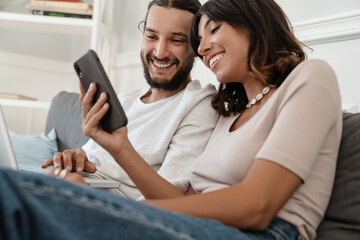 Photo of joyful couple laughing while using laptop and mobile phone