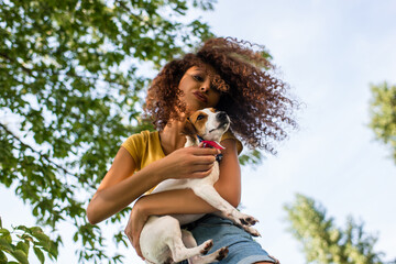 low angle view of curly woman holding jack russell terrier dog and making duck face against blue sky and branches