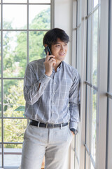 A young Asian businessman stands on the balcony of a window talking on the phone.