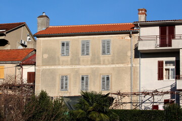 Partially renovated attached large old family house with old dilapidated facade and closed cracked wooden blinds and new roof tiles with shiny new gutter surrounded with other houses and small vegetat