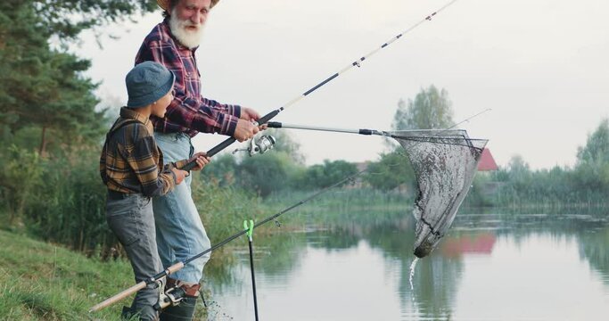 Likable positive senior bearded fishman fishing together with his 10-aged joyful handsome grandchild using fishing rods and net on the pond's bank at daytime in the summer