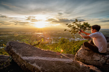man sitting relax and holding a cellphone taking pictures on top of a moutain watching the sunset