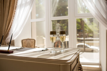 Two glasses of champagne are on the table in a bright room. Wine glasses are in front of the window. Wedding glasses