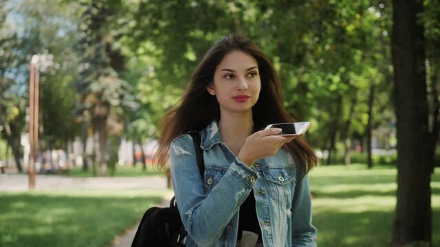 Happy girl activating virtual assistant on smartphone. Smiling young student recording audio message in social network for friend or parents by gadget outdoor on the campus.