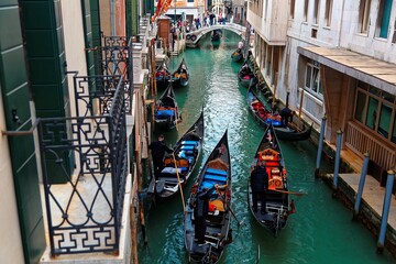 Top view from a balcony over a narrow canal in romantic Venice, with gondoliers steering gondolas through the waterway flanked by old colorful houses & tourists walking on an arch bridge in background