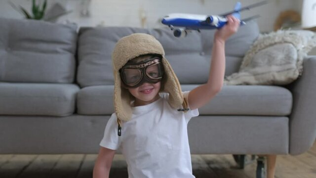 Boy dreams to learn to fly. Happy little boy child playing with white passenger toy plane in glasses and pilot's hat, sitting on floor of an apartment during day. Concept of travel and adventure.