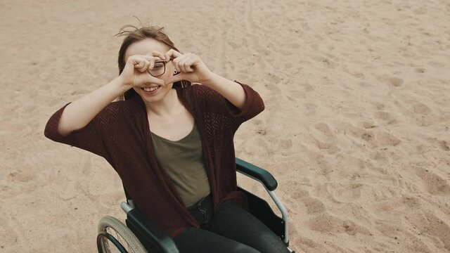 Suport for disabled people. Young woman in the wheelchair on the sandy river coast showing heart with hands. High quality 4k footage