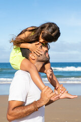 Sweet little girl kissing her dad while riding on his neck. Father and daughter walking or standing on beach by sea. Medium shot. Family leisure time concept