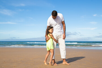 Sweet little girl hugging her dads leg while they walking on golden sand by ocean. Full length. Parenting and family leisure time on beach concept