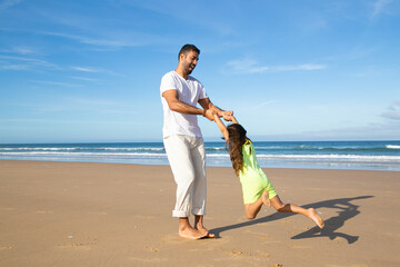 Joyful handsome dad having fun with daughter on beach, playing active games, holding girls hands...