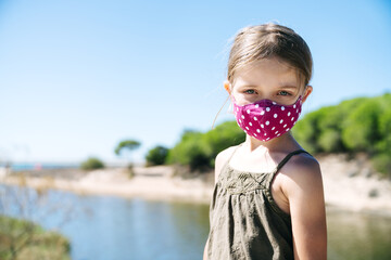 portrait of a blonde girl with blue eyes wearing a face mask on vacation on a beach with pine trees...