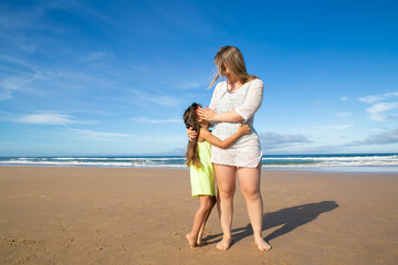 Happy young mom and cute black haired girl hugging while standing on ocean beach. Front view, full length. Motherhood or vacation by sea concept