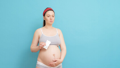 sad young pregnant woman on blue background,  holding condom in her hands