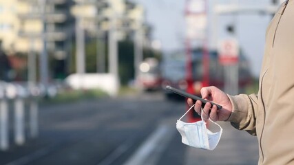 Young man with face mask in hand using smart phone during waiting for tram. Themes modern technology in new normal, coronavirus and personal protection.  - Powered by Adobe